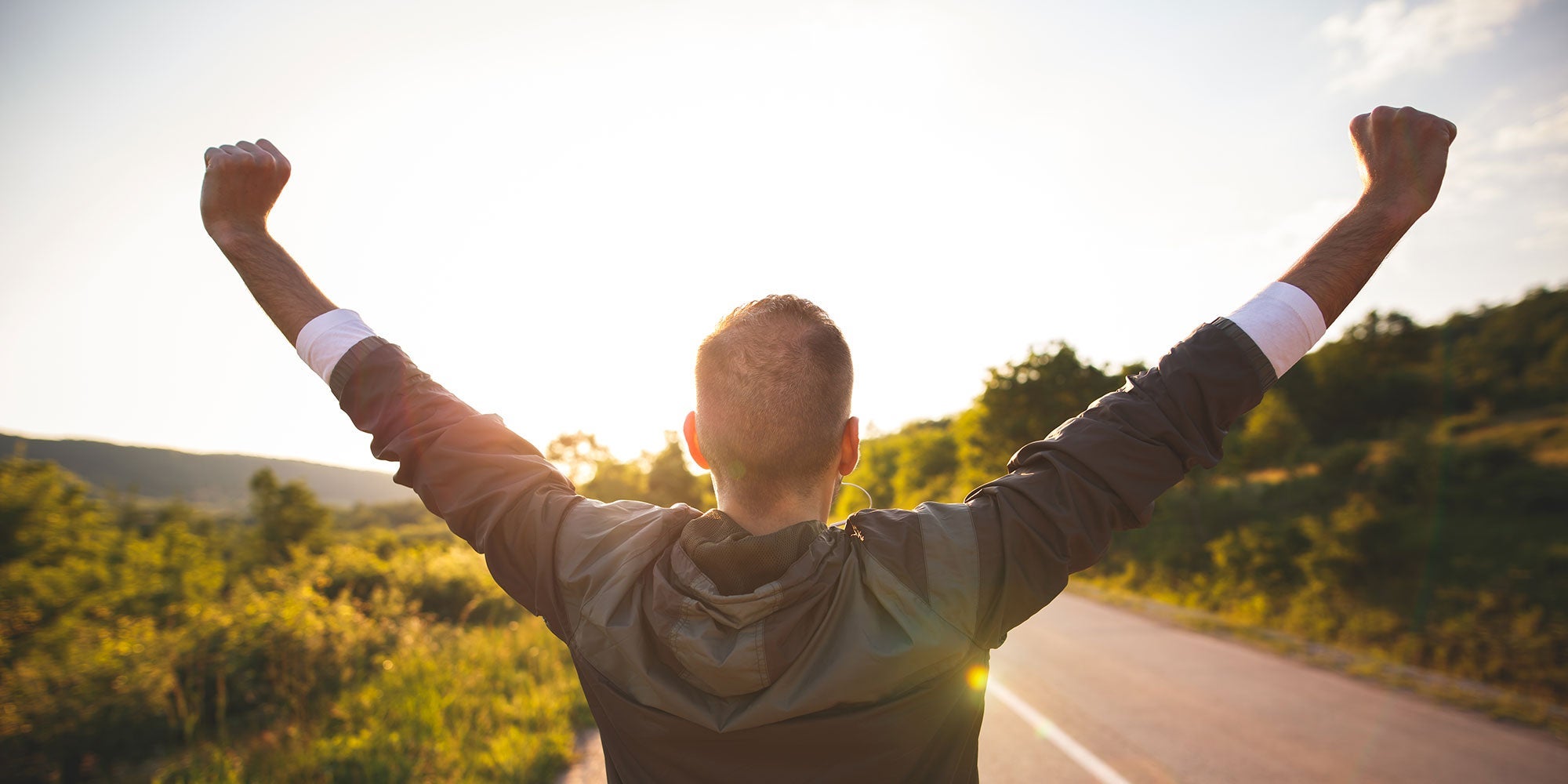 A man stands with the back to the camera, raising his arms triumphantly after finishing a hike