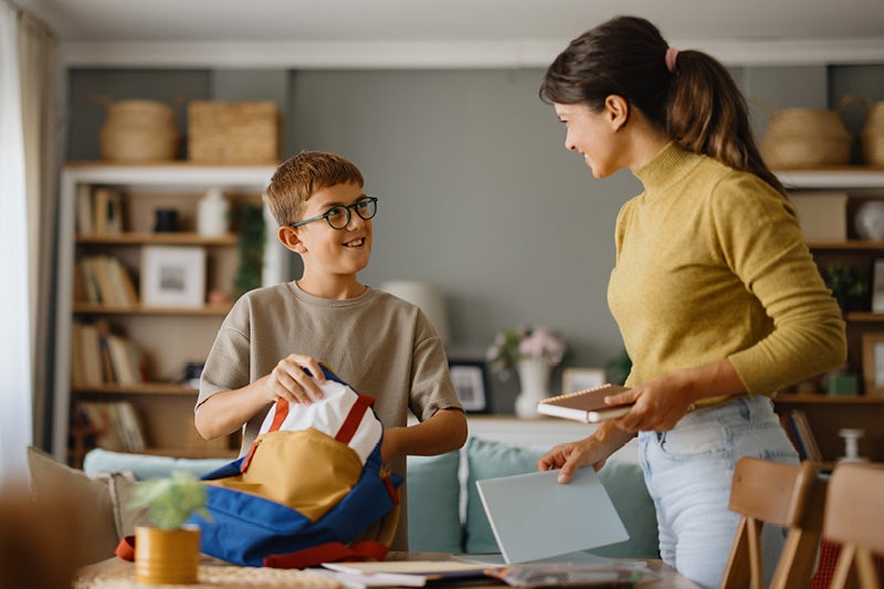 A boy packing a backpack next to a woman with a notebook